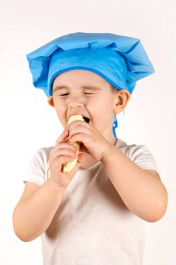 Aşçı şapkalı küçük bir çocuk oyuncak mutfakta yemek pişiriyor ve beyaz arka planda muz yiyor.