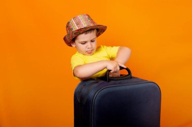 Panama şapkalı ya da şapkalı güzel mutlu bir çocuk turuncu bir arka planda mavi bavulla seyahat ediyor.
