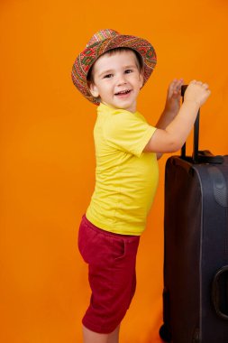 Panama şapkalı ya da şapkalı güzel mutlu bir çocuk turuncu bir arka planda mavi bavulla seyahat ediyor.