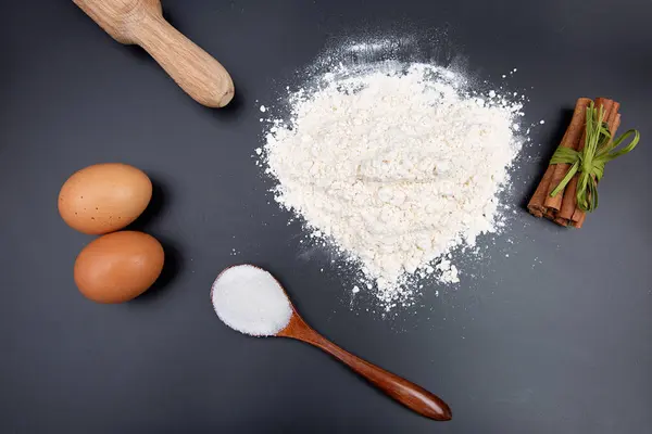 Fırın malzemeleri: bir kase ya da ahşap tabakta buğday unu, kahverengi yumurta ve siyah arka planda bir merdane. Yukarıdan görünüşü.