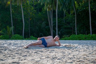 Kumsalda eriyen genç bir adam kumların üzerinde uzanıyor ve güneşleniyor. Dinlenen bir turist poz verir. Egzotik beyaz kum manzarası Andamand deniz plajı ve palmiye ağaçları Güneydoğu Asya, Tayland.