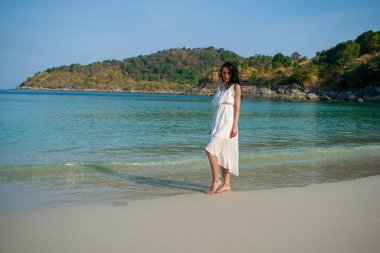 Uzun beyaz elbiseli genç, romantik bir esmer kız kumda durup poz veriyor. Denizde dinlenen bir turist. 