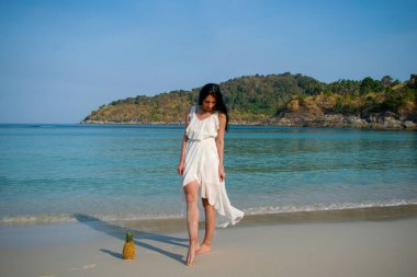 Uzun beyaz elbiseli genç, romantik bir esmer kız kumda duruyor ve yanında meyve ananası ile poz veriyor. Dinlenen bir turist denizde poz verir..