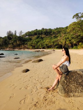 Genç, romantik esmer bir kadın bir taşın üzerine oturur, rahatlar, meditasyon yapar ve gün batımını seyreder. Dinlenen bir turist. Egzotik tropikal Andamand sahili, palmiye ağaçları, dağlar..