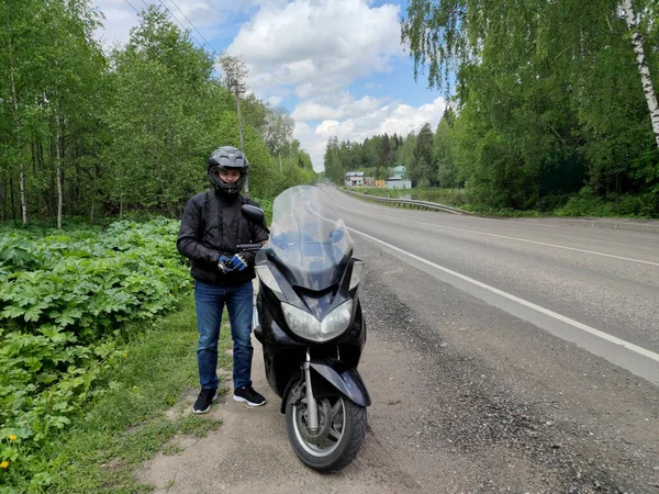 一个骑摩托车的年轻人穿着黑色防护头盔和夹克站在路边的摩托车旁边 森林里阳光灿烂的一天 — 图库照片