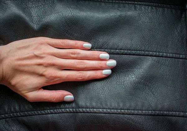 Manicure with shiny pearl nail polish, light color. Powder for design. Holographic hue. Woman hand with a pastel manicure on a black leather background.