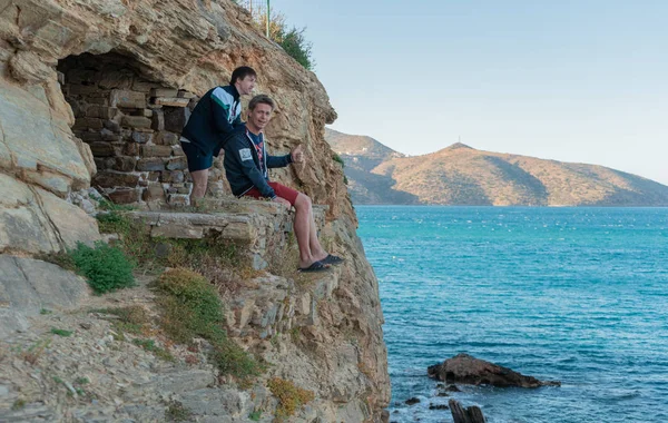 two men sit on the edge of a cliff and look at the sea