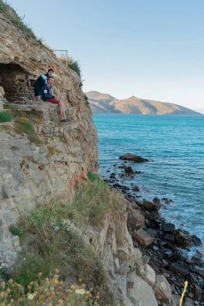 two men sit on the edge of a cliff and look at the sea