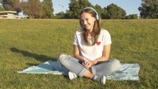 Sorrindo jovem com assento de fones de ouvido sem fio na grama verde — Vídeo de Stock