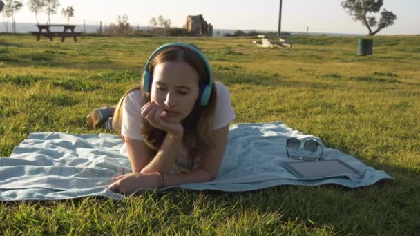 Kvinna låg på gräset i parken och lyssna på musik i hörlurar — Stockvideo