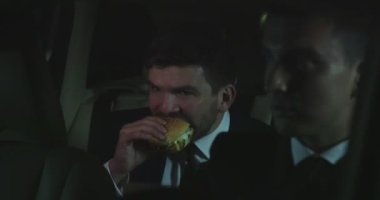 Adam bir arabanın arka bir burger yemek.