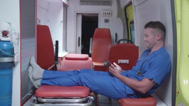 Чоловік сидить з телефоном у машині швидкої допомоги, дивиться щось і посміхається . — стокове відео