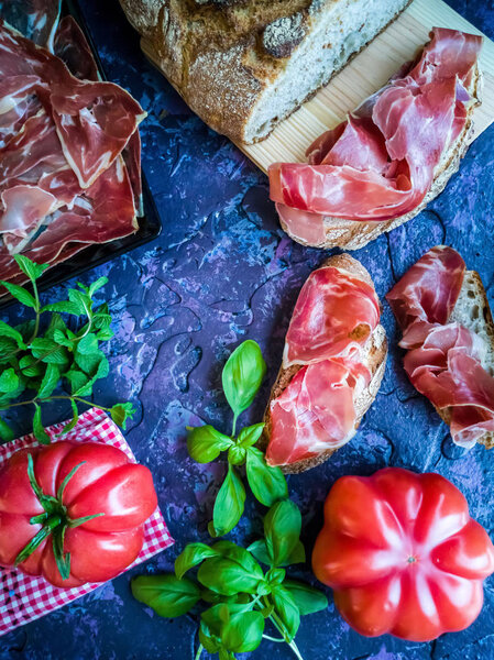 Ham, tomato, bread and herbs in composition on dark background with rough texture