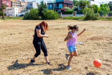 Anne ve kızı tarlada portakal topuyla top oynuyorlar. Kuru otlarla. Koronavirüsü önlemek için maske takıyorlar. Sağlıklı yaşam konsepti
