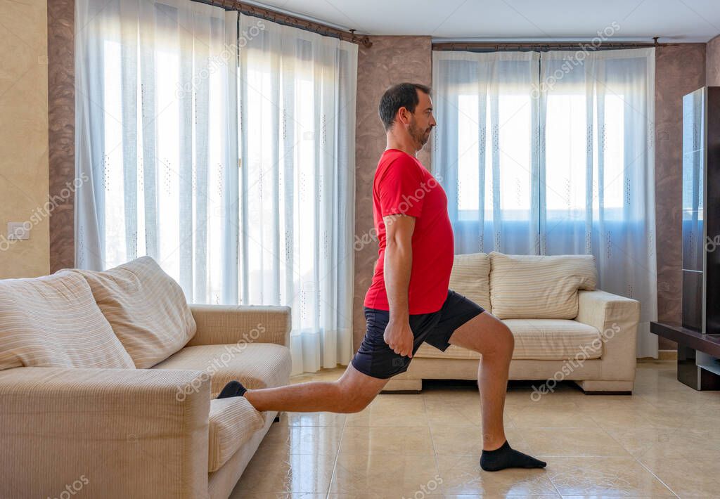 Bearded man in low shape exercising with black and red sportswear in his living room in front of the sofas on a mat