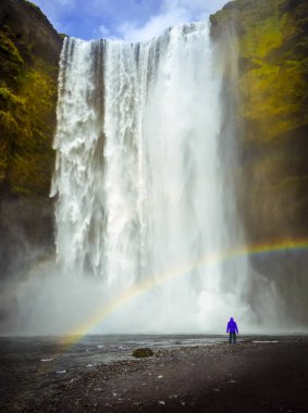 İzlanda 'da Skogafoss şelalesi. Mavi ceketli adam şelaleye bakıyor. Gökkuşağı