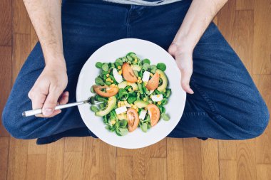 Kot pantolonlu genç adam otururken sebzeli lezzetli salata yiyor.