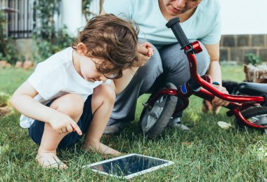 A young boy with his mother sitting on the grass in a park using a tablet PC. Technology, lifestyle, education, people concept. clipart