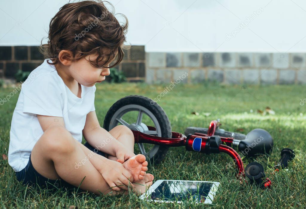 A little boy sitting on the green grass and looking at a digital tablet with a red bike.