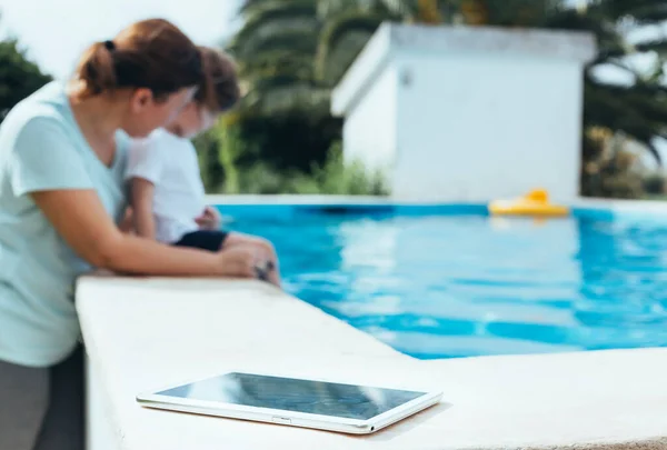 Digital Tablet photo in the foreground with mother and son playing in the pool in the background. Concept of the digital switch-off. Selective focus.