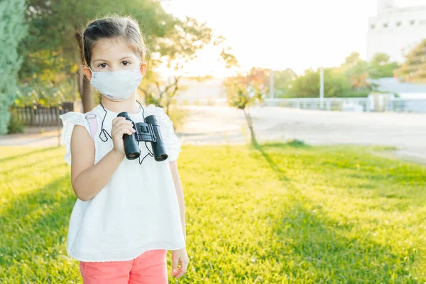 带着面具和背包的女孩在公园里玩望远镜 Coronavirus — 图库照片