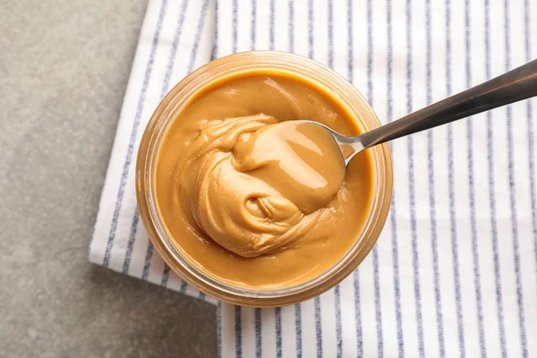 Glass jar with peanut butter and spoon, and kitchen towel on gre — Stock Photo, Image