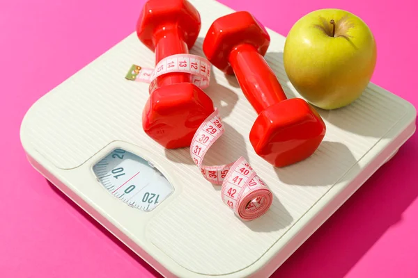 Весовые весы, яблоко, гантели и измерительная лента на розовом backgr — стоковое фото