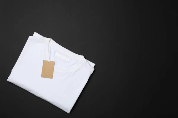 Folded blank white t-shirt with tag on black background, space f — Stock Photo, Image