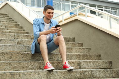 Genç adam merdivenlerde oturur ve telefona bakar.