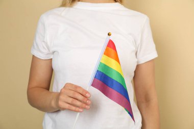 Bej arka planda LGBT bayrağı taşıyan beyaz tişörtlü genç bir kadın.