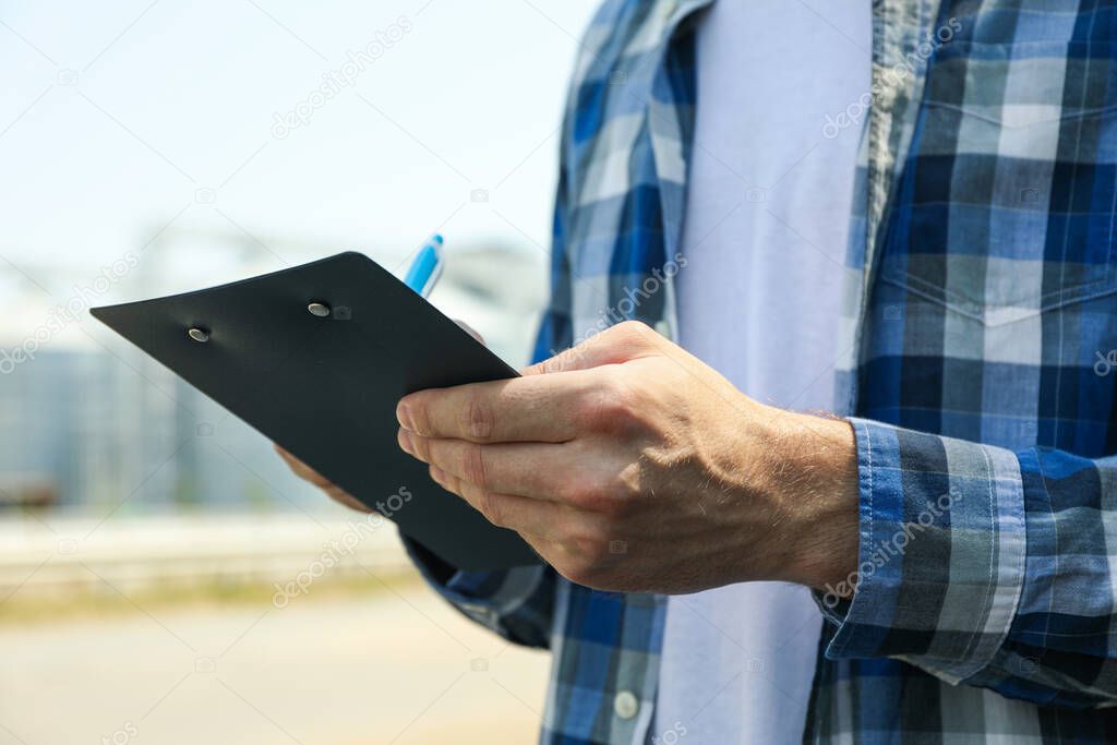 Young man with clipboard and pen against grain silos. Agriculture business