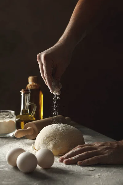 Man sprinkle flour on dough against dark background