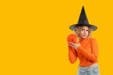 Turuncu giyinmiş bir cadı balkabağını parlak arka planda tutar.