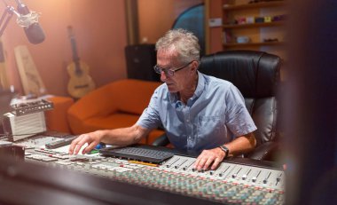 Male middle aged recording engineer in a recording studio sitting at a multi channel console clipart