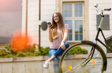 Şehirde bisiklet sürerken akıllı telefon kullanan çekici genç bir kadın.