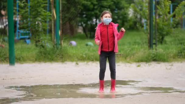 医者のマスクとゴムブーツの女の子は雨の後の春の日に水たまりを通ってジャンプします Covid 19コロノウイルス感染からの保護の概念 — ストック動画