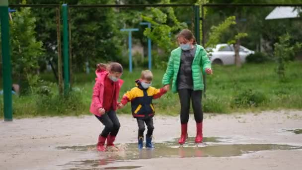 Dwie Dziewczynki Chłopiec Maskach Medycznych Gumowych Butach Skaczą Przez Kałuże — Wideo stockowe