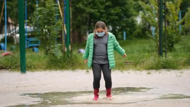 医者のマスクとゴムブーツの女の子は雨の後の春の日に水たまりを通ってジャンプします Covid 19コロノウイルス感染からの保護の概念 — ストック動画