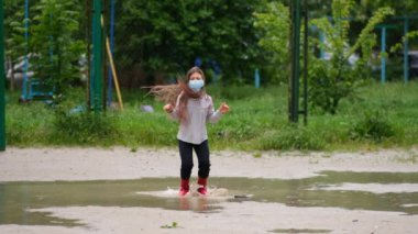 Tıbbi maskeli ve lastik çizmeli küçük bir kız yağmurdan sonraki bahar günü su birikintilerinden atlıyor. Covid-19 koronovirüs enfeksiyonundan korunma kavramı.