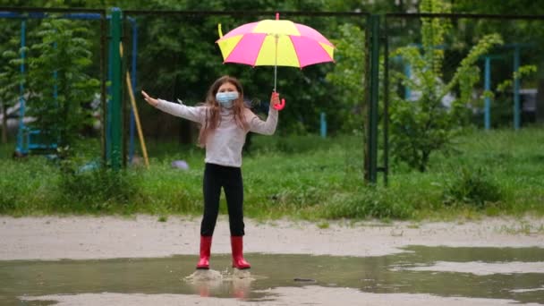 医療用マスクとゴム製のブーツの傘を持つ少女は 雨の後の春の日に水たまりを通ってジャンプします Covid 19コロノウイルス感染からの保護の概念 — ストック動画