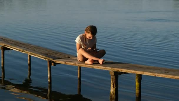 スマートフォンを持った女の子が夕暮れ時に湖の上の木製の桟橋に座っています 夏の屋外レクリエーション 幸せな子供時代の概念 — ストック動画