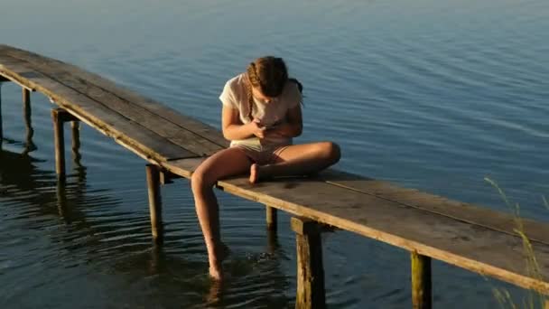 スマートフォンを持った女の子が夕暮れ時に湖の上の木製の桟橋に座っています 夏の屋外レクリエーション 幸せな子供時代の概念 — ストック動画