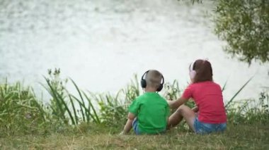 Küçük bir oğlan ve kız nehir kıyısında kulaklıklarla oturup müzik dinliyorlar. Açık hava eğlencesi kavramı.