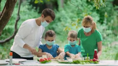 Anne ve baba çocuklara ızgara et pişirmeyi öğretiyor. Tıbbi maskeli bir aile doğada piknik yapıyor. Covid-19 salgını sırasında dinlenme.