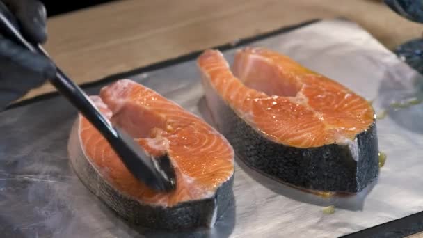 A chef in black gloves is using a cooking brush to smear vegetable oil on pieces of red fish for cooking in foil . close up. Preparation of fish. Salmon steak. — Stock Footage Chef Black Gloves Using Cooking Brush Smear Vegetable Oil Pieces Stock Video
