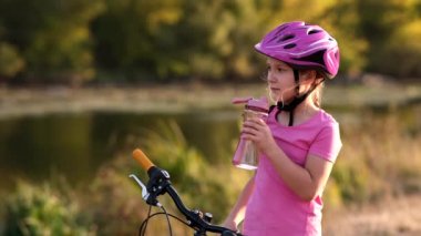 Bisiklet kasklı küçük bir kız nehir kıyısındaki bir bisikletin yanında temiz su içiyor. Açık hava sporları konsepti.