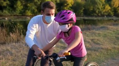 Genç bir baba kızına tıbbi maskeli bisiklet sürmeyi ve nehir kenarında bisiklet kaskı takmayı öğretiyor. Covid-19 koronavirüs enfeksiyonundan korunma. 