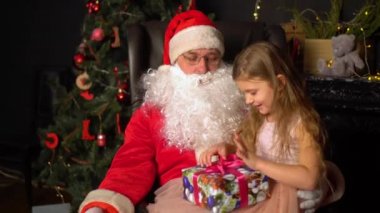 Noel Baba küçük bir kıza hediye verir. Noel ağacı olan bir odada kollarında oturan bir kız..