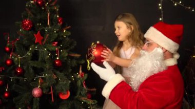 Noel Baba ve küçük bir kız Noel ağacı süsler. Noel Baba kızı kollarına aldı ve büyük kırmızı bir top astı..