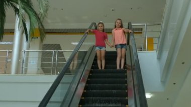 İki küçük kız alışveriş merkezinde yürüyen merdivenden aşağı iniyor.